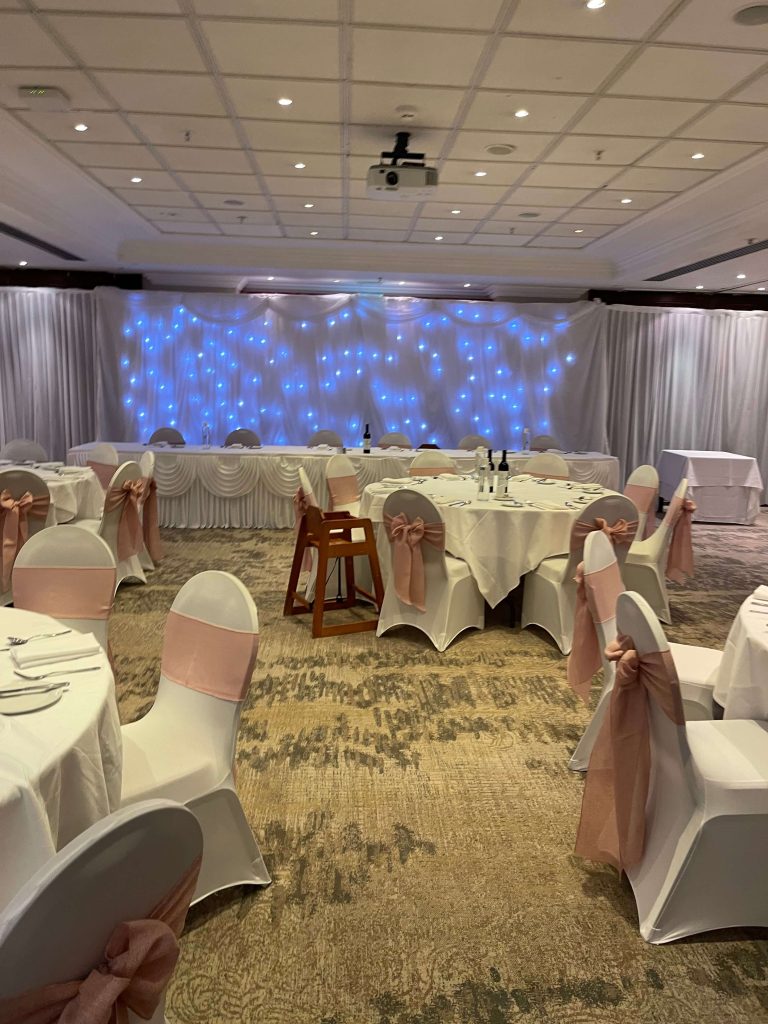 Dressed function room set for a wedding with tables, chair covers with pink sashes, a top table with starlight backdrop behind